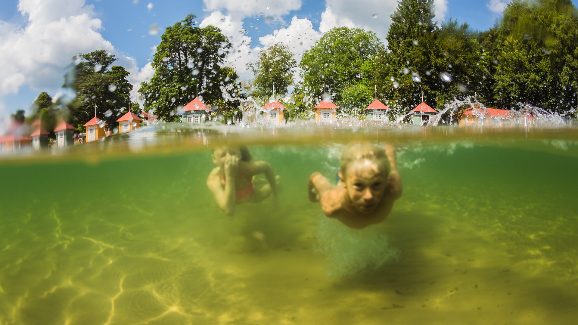 Två barn simmar under vattnet vid stranden på Guldkroksbadet. Badhytterna syns i bakgrunden.