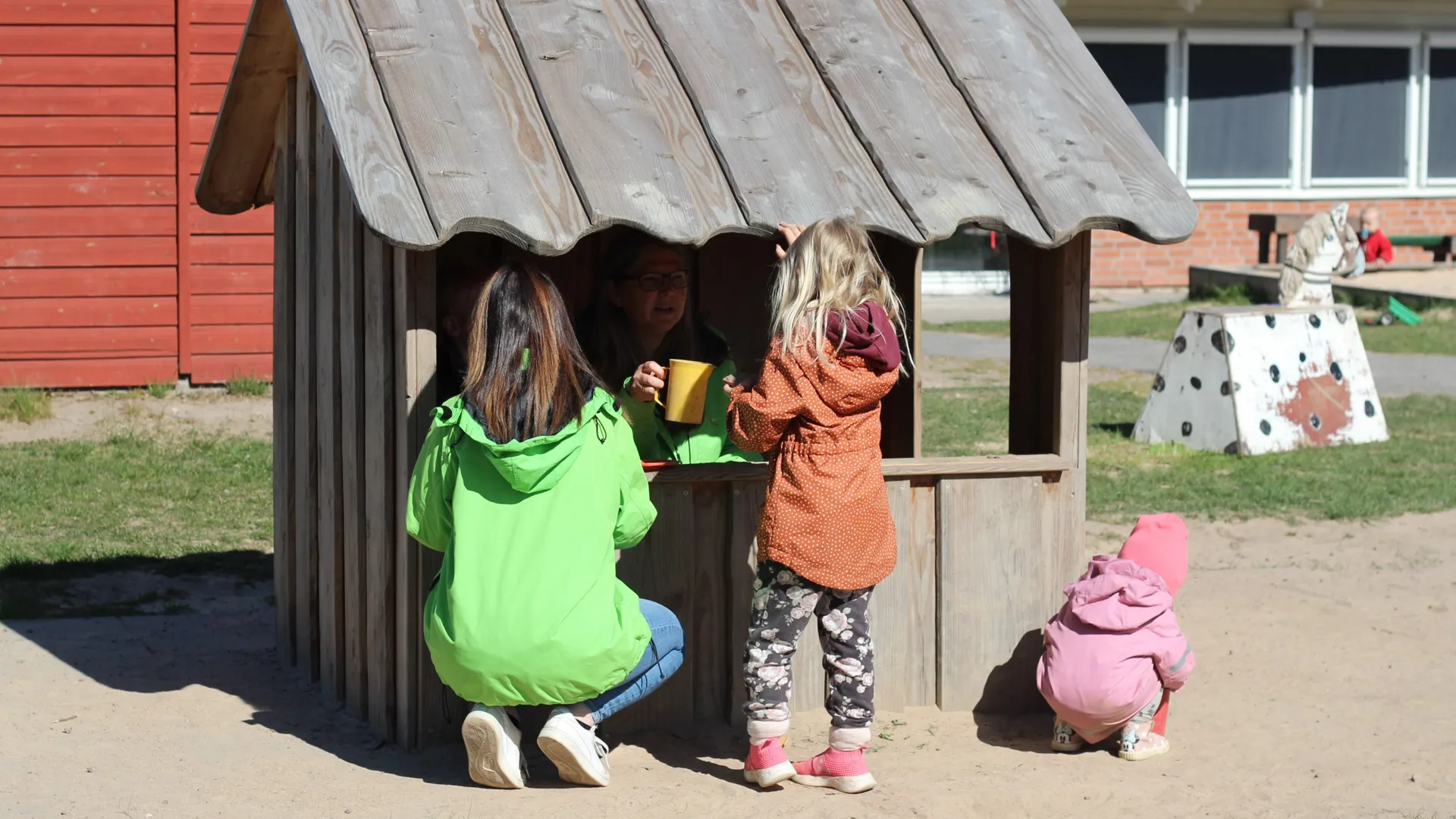 Pedagog och förskolebarn framför ett litet trähus som fungerar som kiosk.