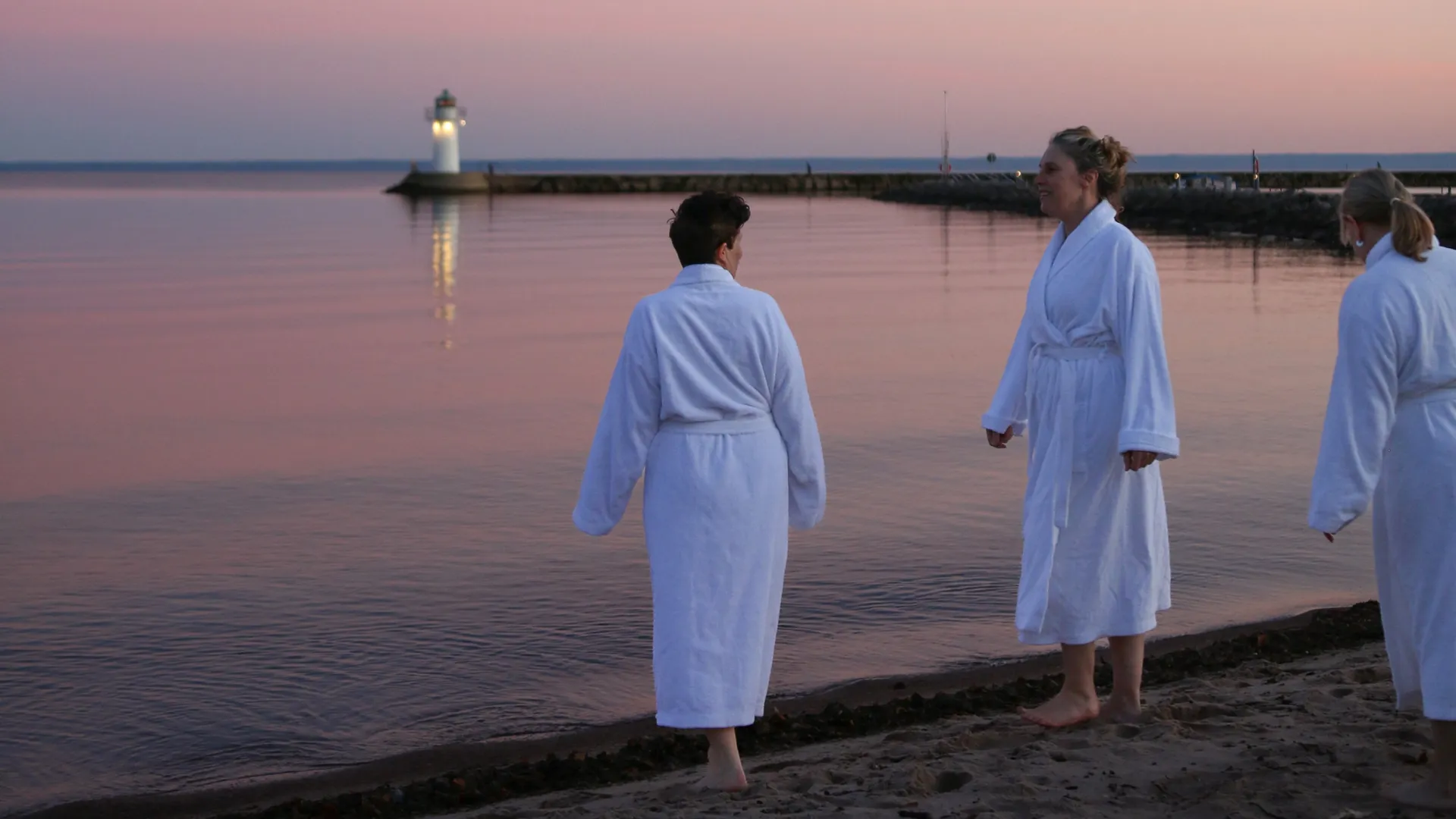 Några människor i badrock står på stranden vid Guldkroksbadet i solnedgången.