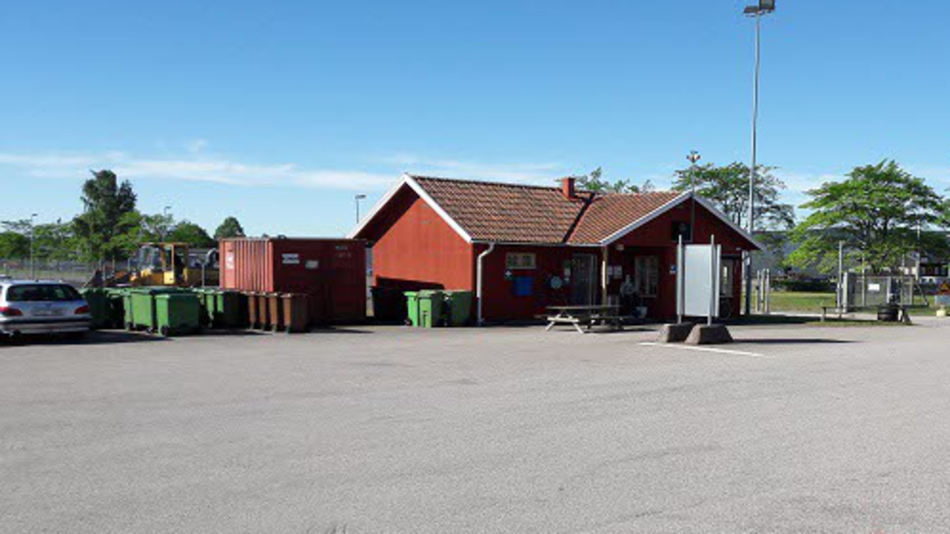 En del av återvinningscentralen i Hjo. Ett rött hus, soptunnor och en container syns i bild.