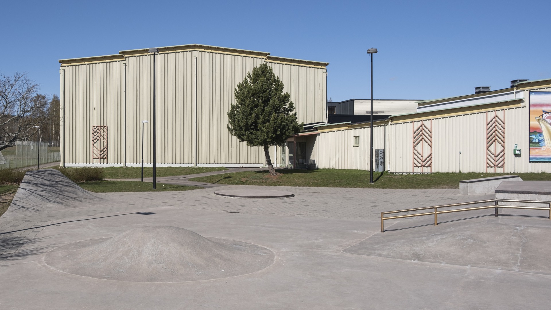 Actionparken vid Guldkrokshallen i Hjo. En grå betongyta med gupp, formationer och räcken som går att åka skateboard eller sparkcykel över. I bakgrunden syns Guldkrokshallen som är en ljusgul byggnad med träfasad.
