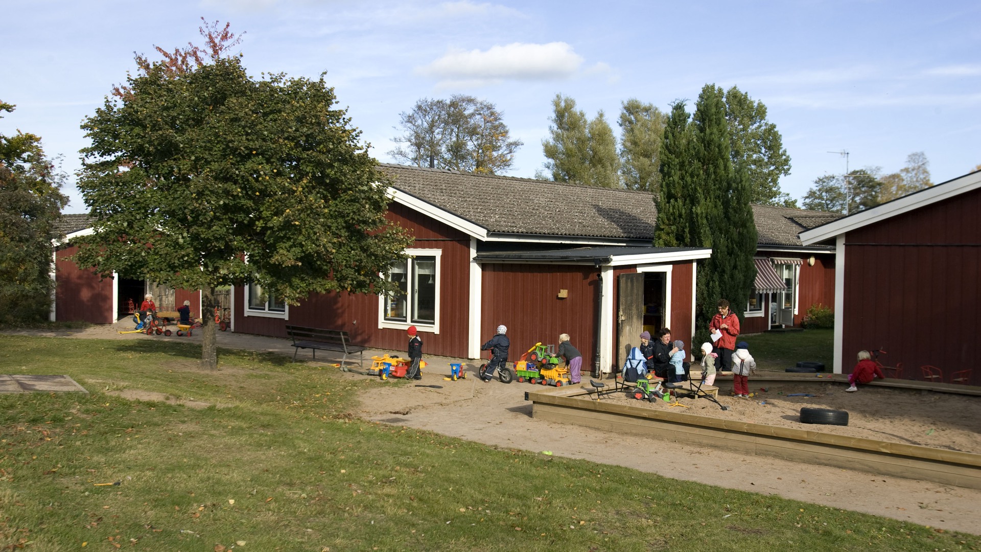 Exteriörbild av Orrelyckans förskola. Röda enplanshus med vita knutar med sandlådor framför.