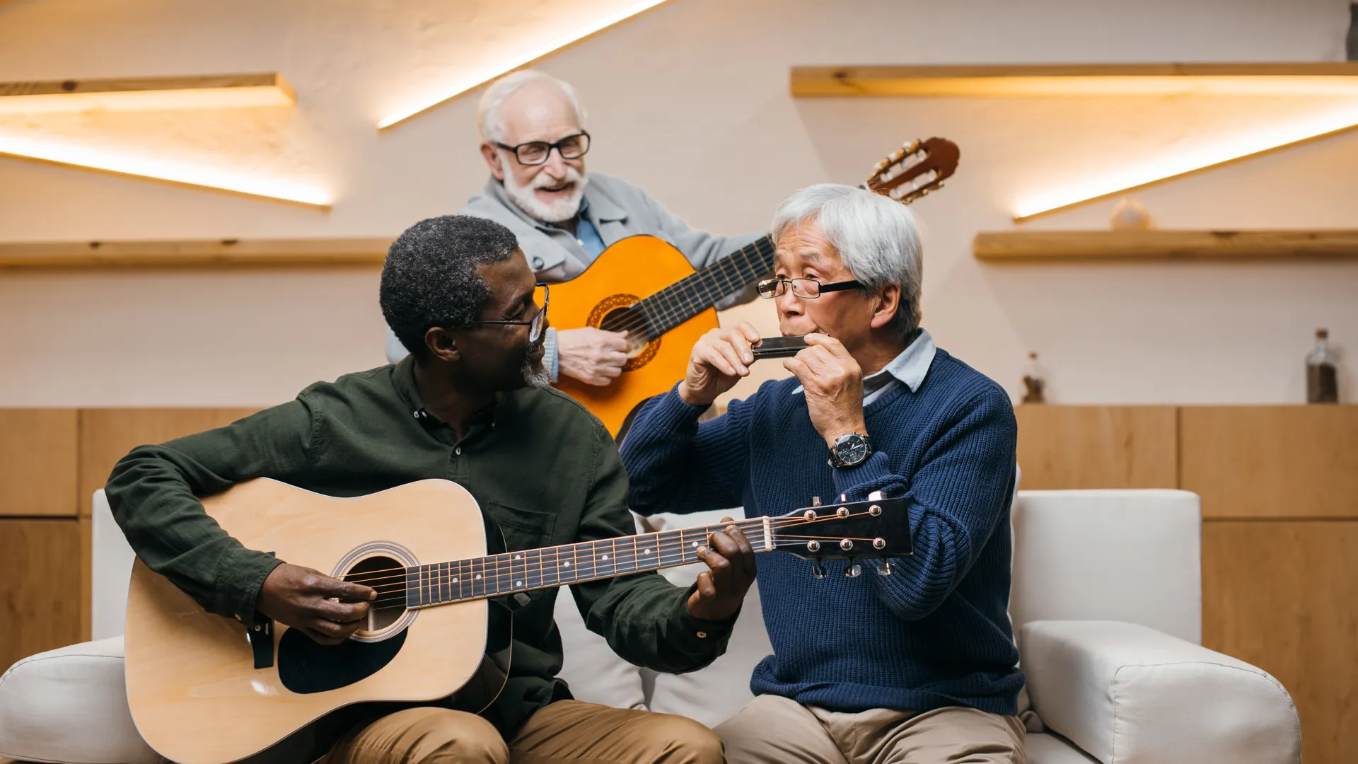 Tre äldre män som spelar musik tillsammans på en soffa med två gitarrer och ett munspel.