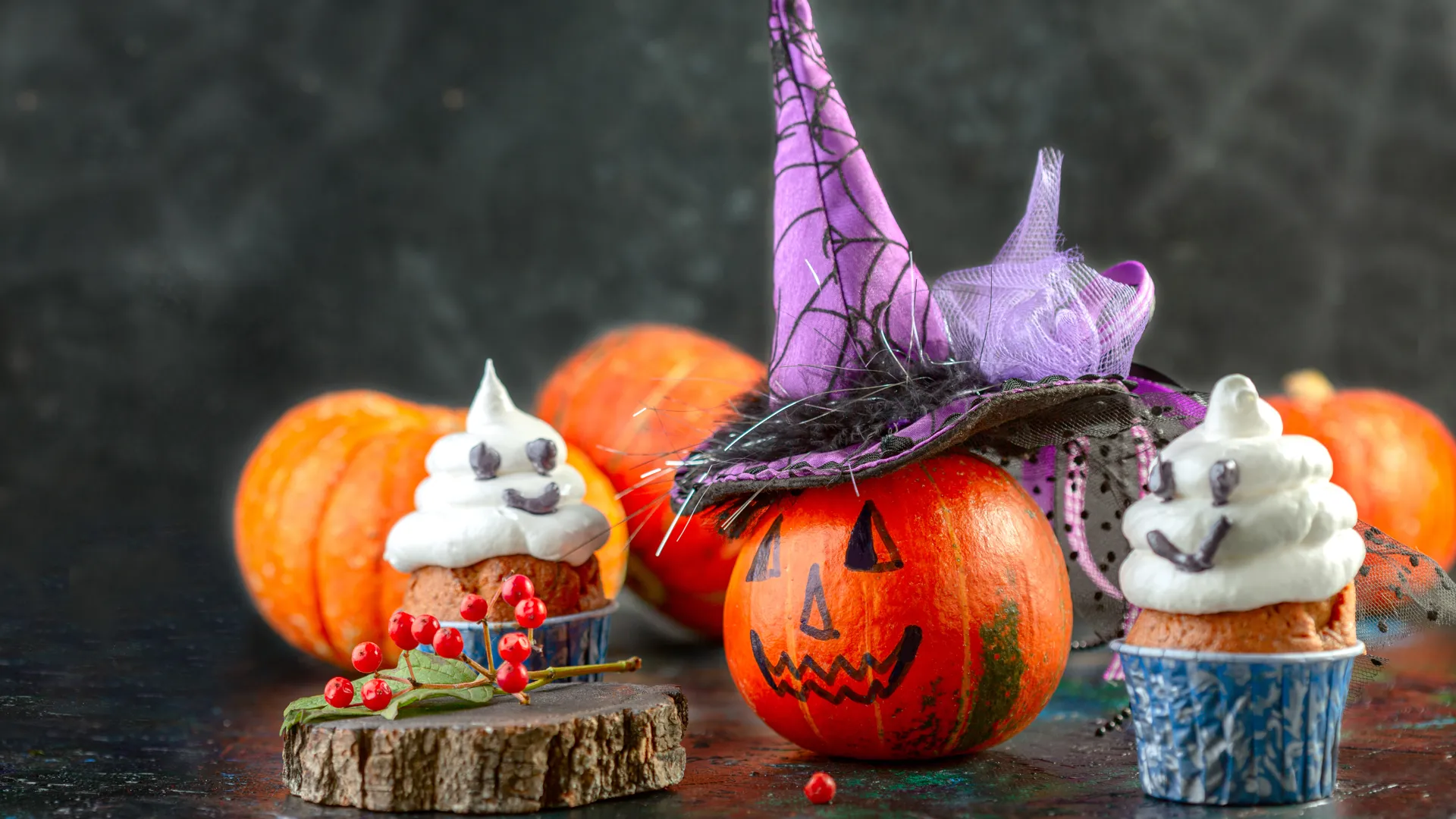 bild med halloween-tema. I bakgrunden ligger ett antal pumpor som är oskarpa. I förgrunden ser man en pumpa, målad med ögon, näsa och mun. Pumpan kröns av en spetsig häxhatt. På bägge sidor om pumpan står muffins med glasyr, dekorerade med ögon och mun.