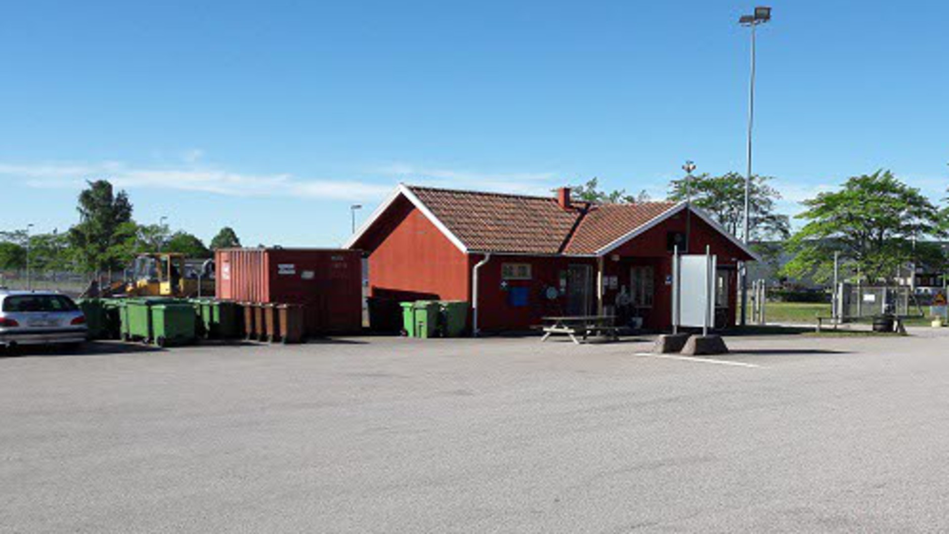 En del av återvinningscentralen i Hjo. Ett rött hus, soptunnor och en container syns i bild.