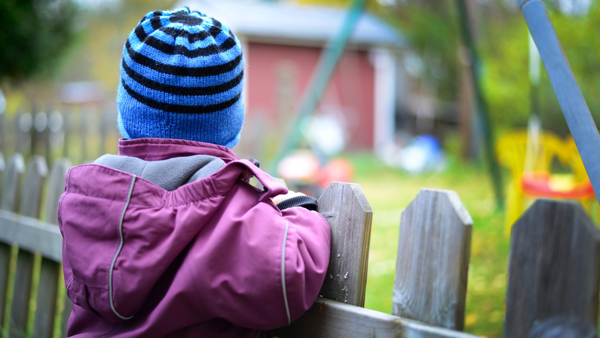 Ett barn i mössa står vid ett staket och tittar bortåt.