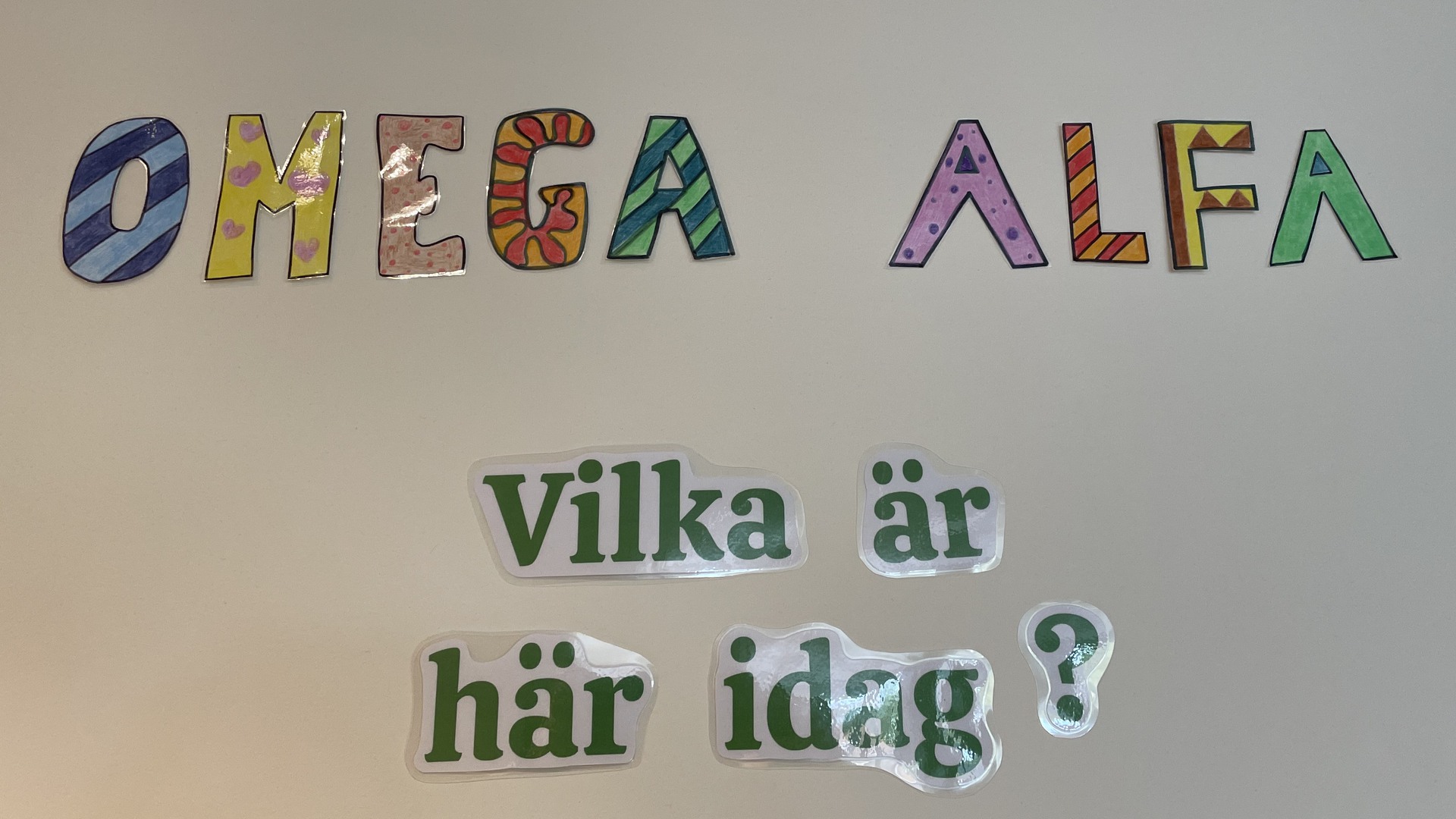 Orden Omega, Alfa och frasen Vilka är här idag? sitter uppklistrade på en vit vägg.