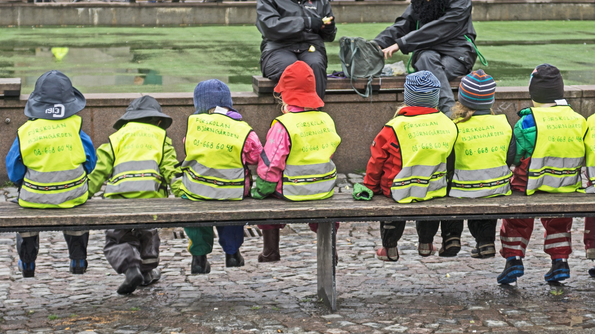 En barngrupp är på utflykt och sitter uppradade på en bänk i reflexvästar.