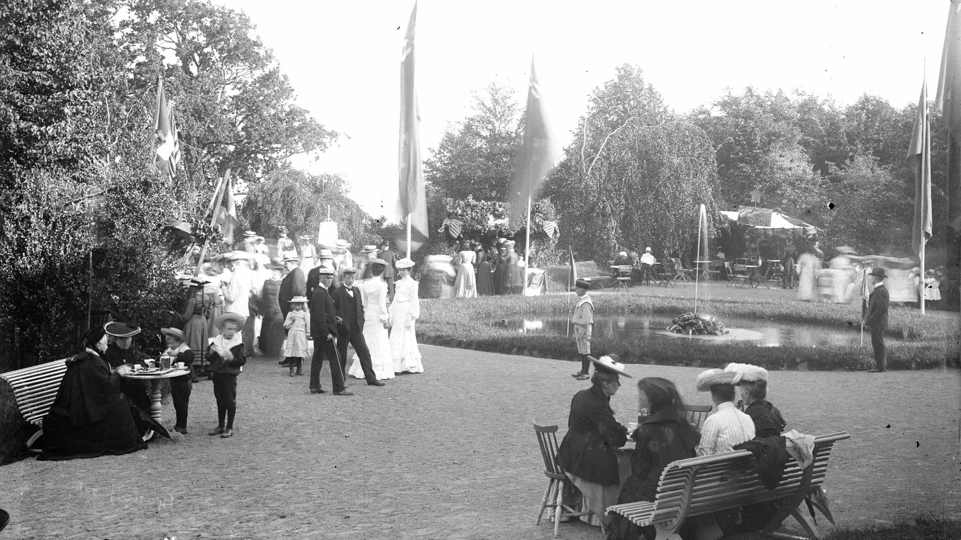 En svartvit, äldre bild från Hjo stadspark. Finklädda personer runt en rund damm.
