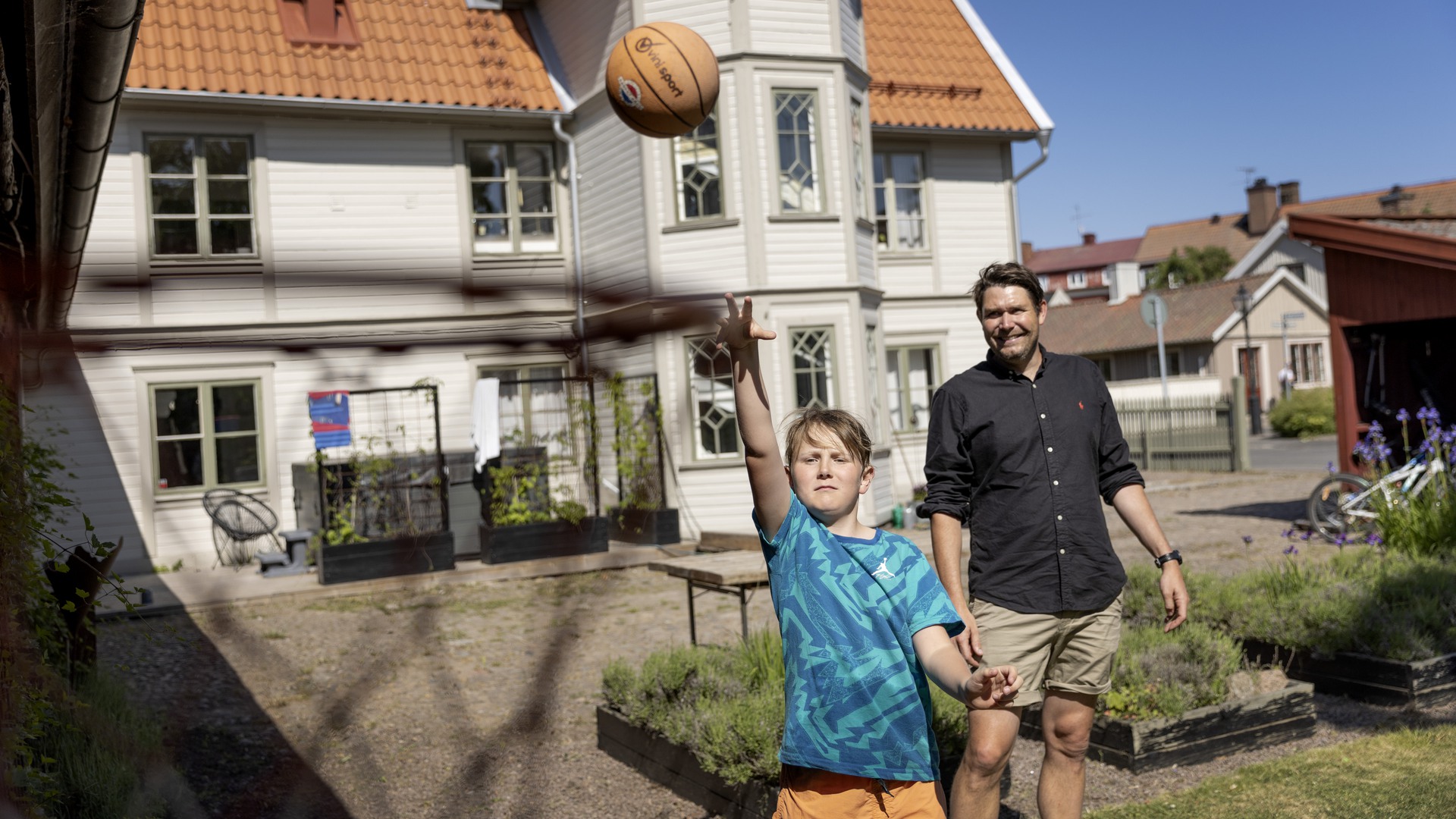 Far och son spelar basket i trädgården, med en stor trävilla i bakgrunden.