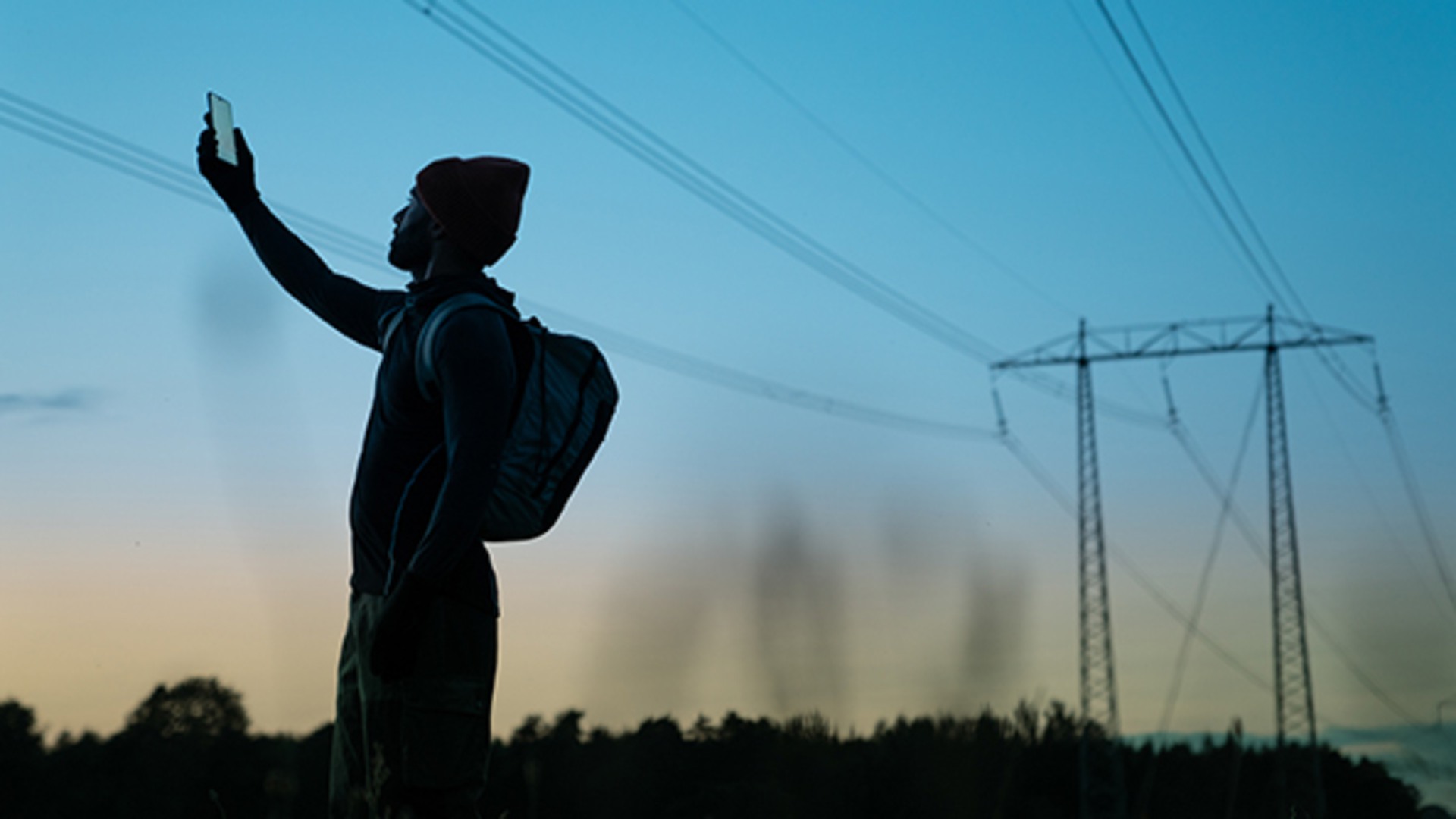 Man söker mobiltäckning genom att lyfta mobilen upp i luften. I bakgrunden syns elledningar och skog i mörk siluett.