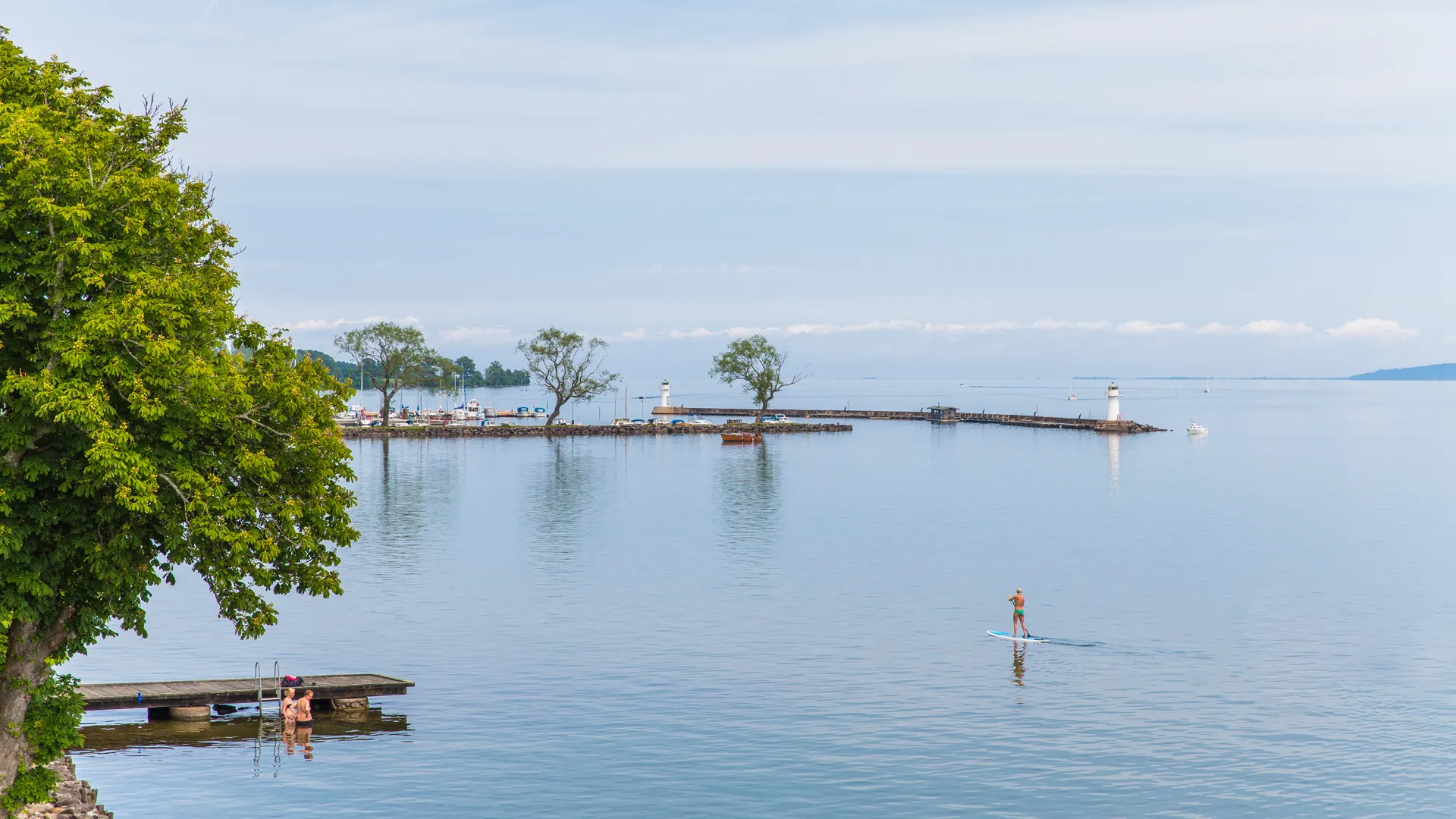 Flygbild från strandpromanden, ut över Vättern. Fyren, vågbrytaren, en SUP-bräda och några som badar.