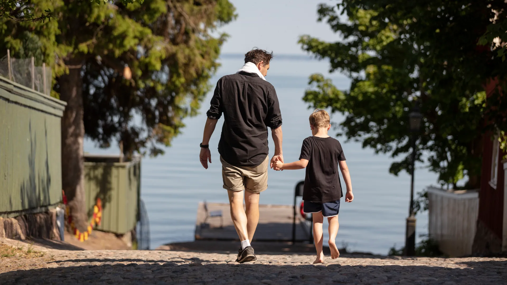 En vuxen och ett barn håller varandra i handen och promenerar ner mot Skolbryggan i Hjo.