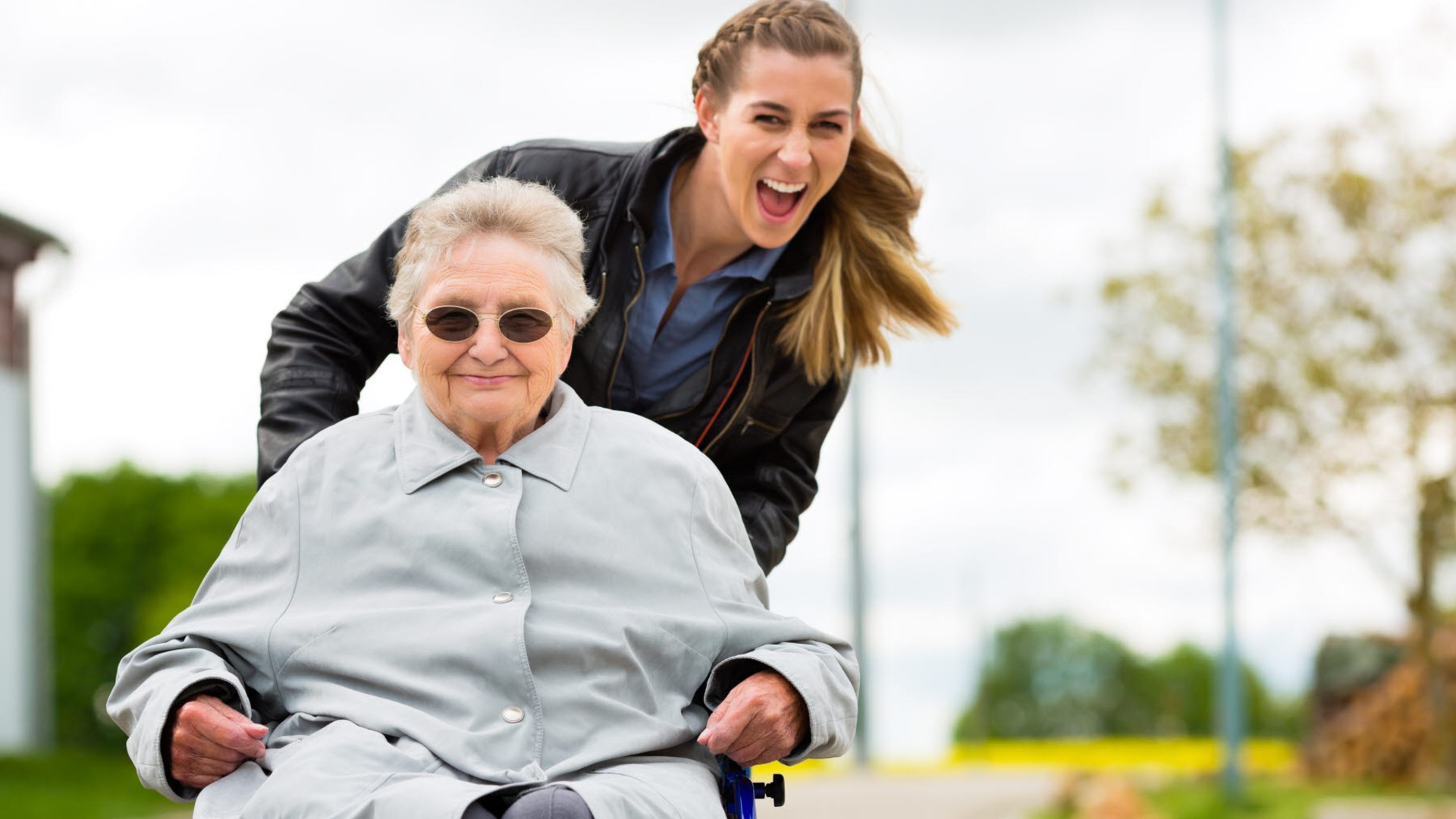 Vårdpersonal kör en äldre person i rullstol. Båda ser väldigt glada ut.