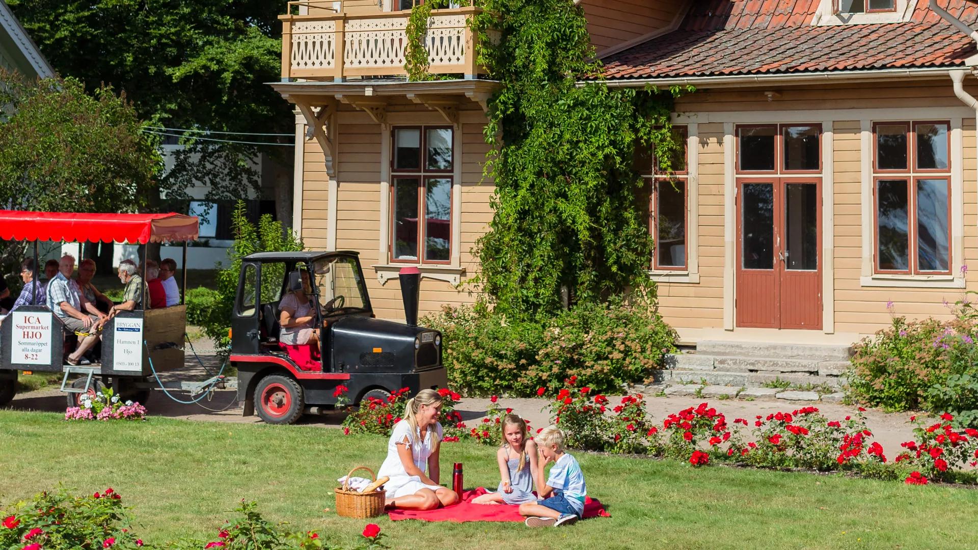 En familj har picknick på gräset i stadsparken. I bakgrunden syns en trävilla och ett turisttåg. 