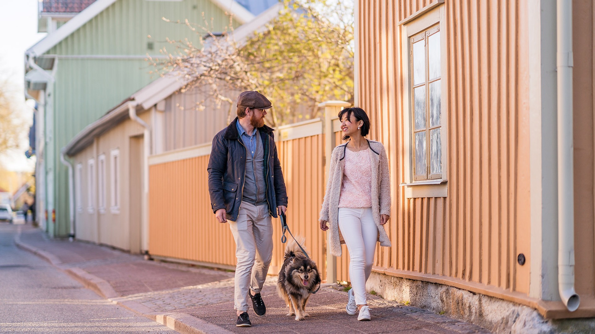 Två personer promenerar med en hund på en gata med trähus.