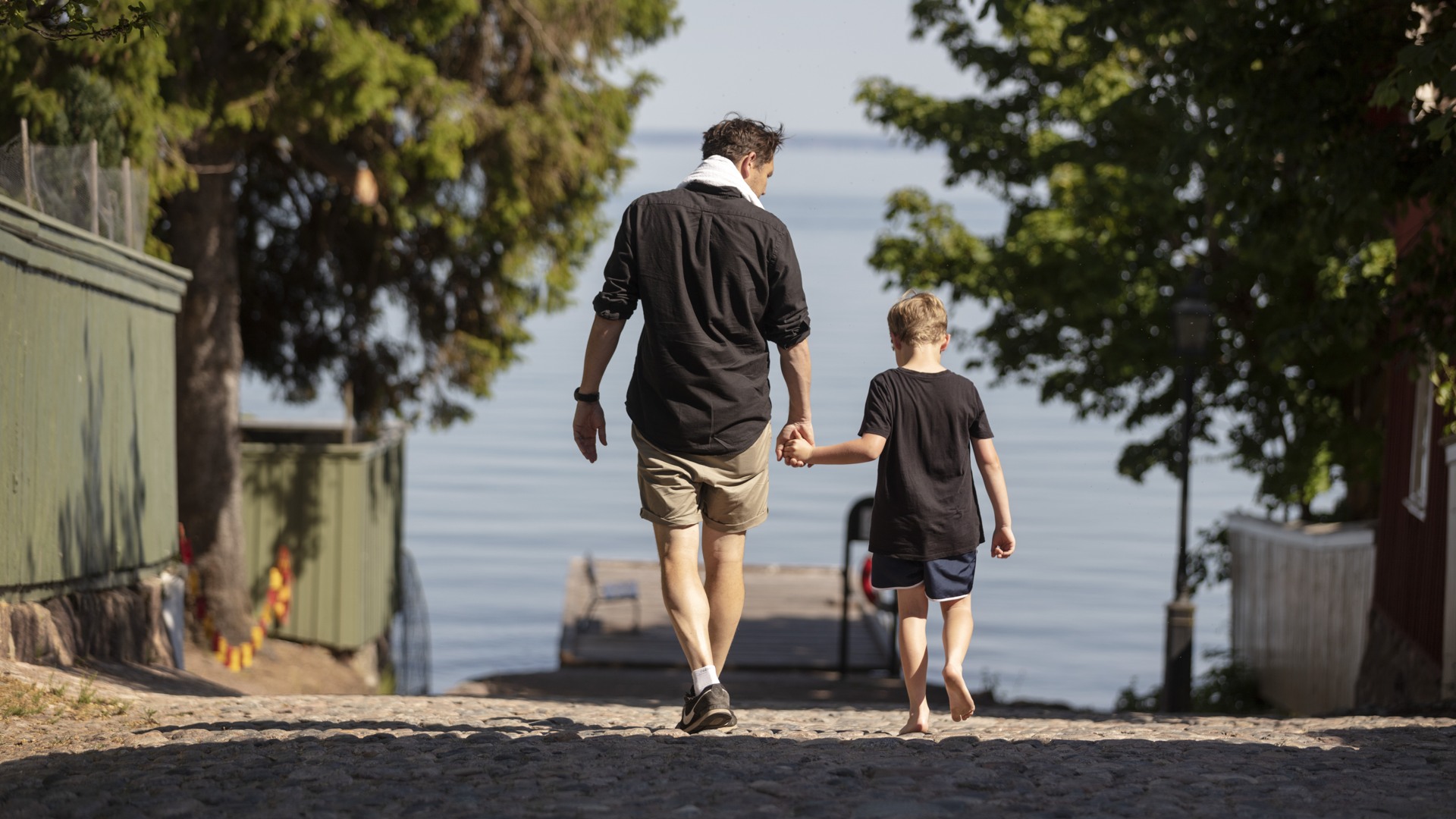 En vuxen och ett barn håller varandra i handen och promenerar ner mot Skolbryggan i Hjo.