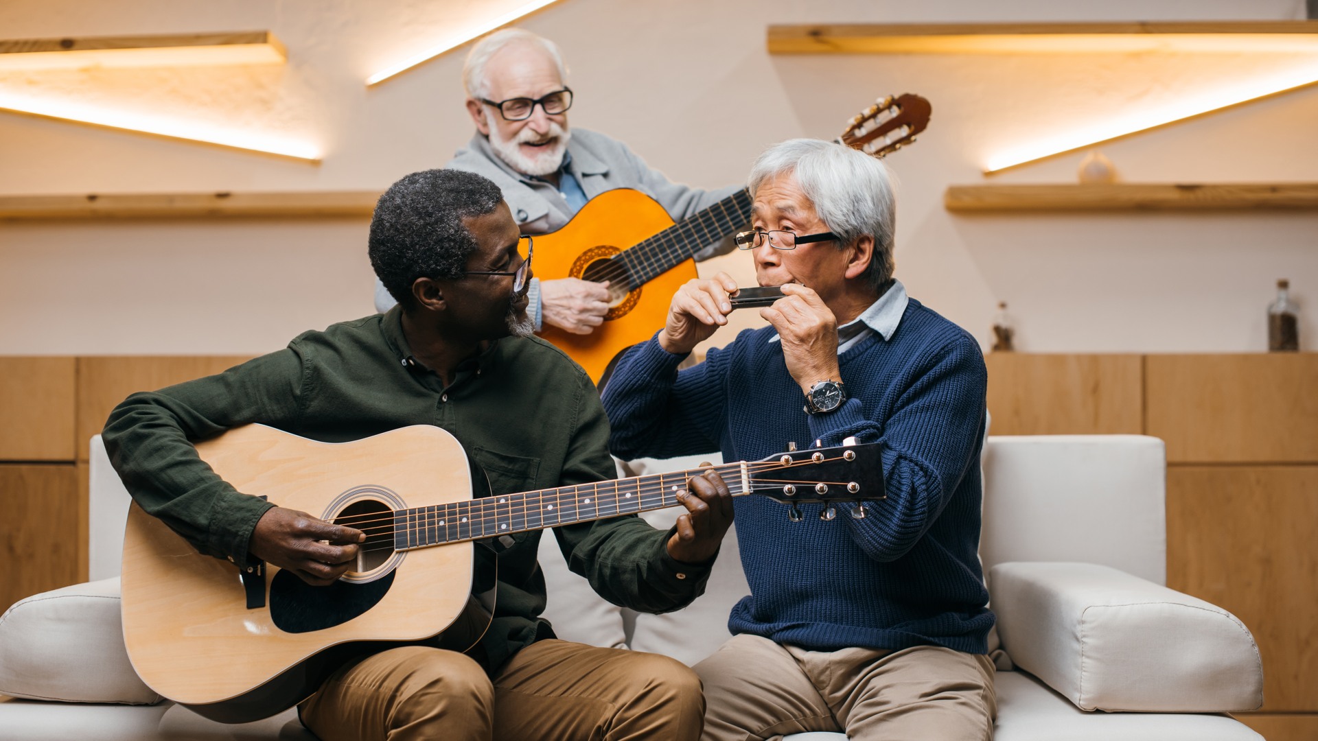 Tre äldre män som spelar musik tillsammans på en soffa med två gitarrer och ett munspel.